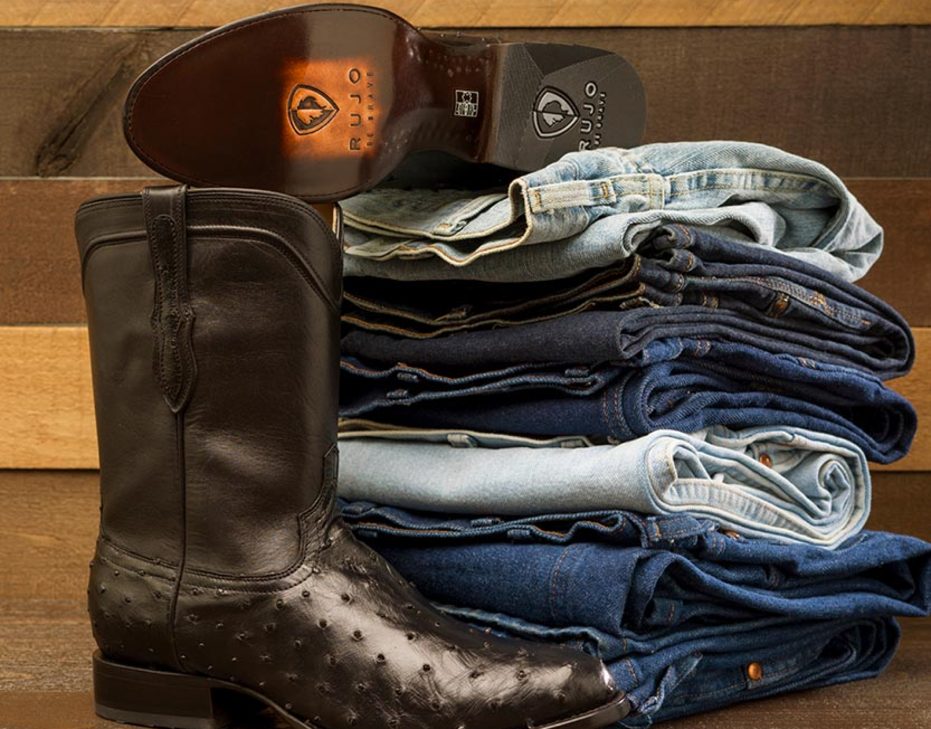 A dark brown ostrich cowboy boot next to a stack of folded blue jeans.